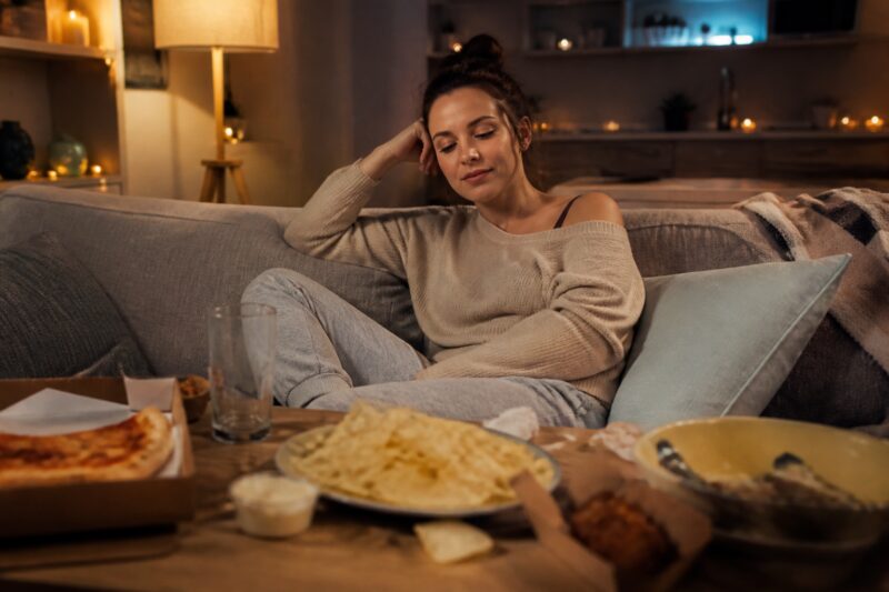 Müde Frau nach Alkoholkonsum, die zu Fast Food auf dem Couchtisch greift, symbolisiert schlechte Ernährungsentscheidungen.