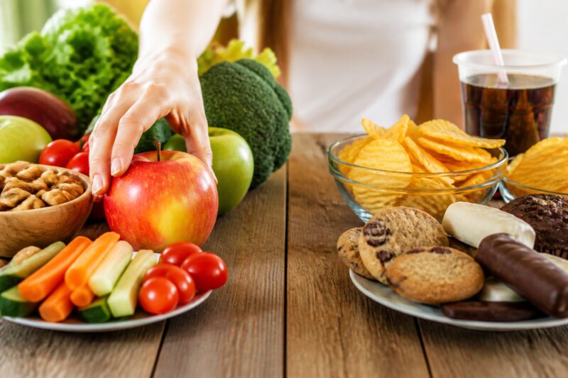 Ein Tisch, auf dem links gesunde Snacks wie Obst und Nüsse und rechts ungesunde Snacks wie Kekse und Chips liegen, mit einer Hand, die nach dem Obst greift.