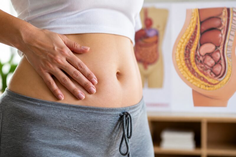 Eine Frauenhand berührt sanft den Bauch, im Hintergrund sind unscharf medizinische Grafiken über viszerales Fett zu erkennen.